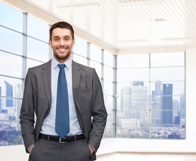 smiling salesperson in a suit with hands in pocket in office with windows with city view